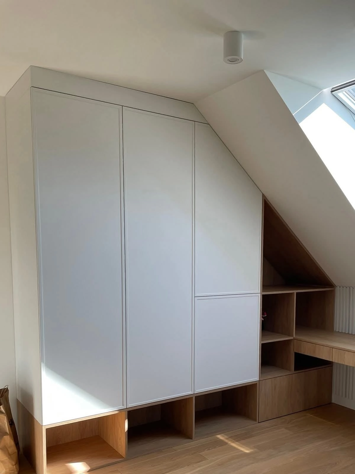 Einbauschrank nach Maß unter einer Dachschräge: grifflose weiße Fronten folgen der Dachneigung, darunter und seitlich offene Fächer und Schubladen in Holzdekor, rechts eine Schreibtischplatte unter dem Dachfenster.