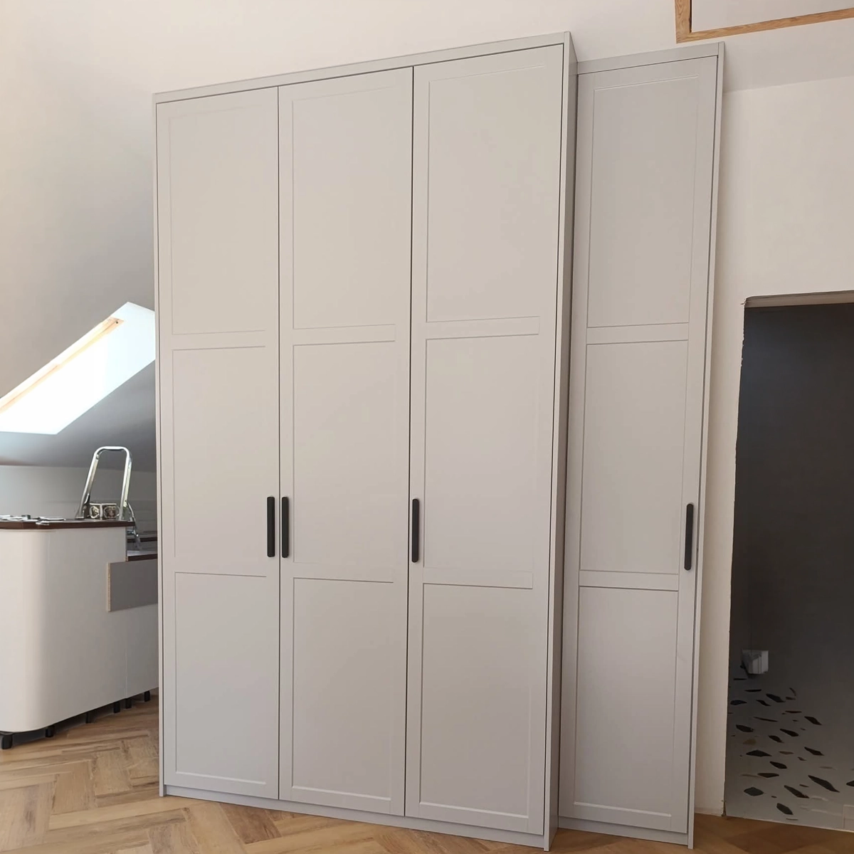 Einbauschrank im Schlafzimmer unter dem Dach: drei breite Türen mit Rahmenfronten in hellem Greige und schwarzen Stangengriffen, daneben ein schmales Zusatzelement, davor Fischgrätparkett.