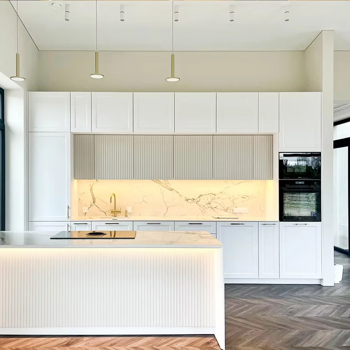 Küche nach Maß mit weißen Kassettenfronten bis unter die Decke, darüber eine Reihe gerillter Oberschränke in Beige; Rückwand und Kücheninsel in Marmoroptik, goldene Armatur und eine Insel mit gerillter Front und indirekter Beleuchtung.